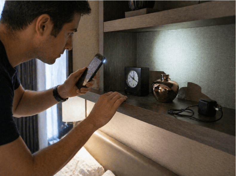 person using a phone flashlight to check a bedroom shelf for hidden cameras