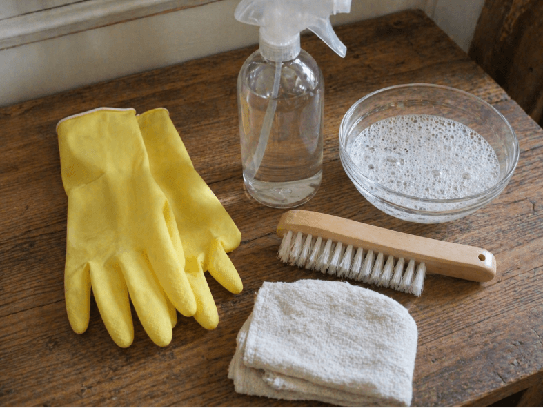 mold cleaning supplies with gloves, spray bottle, brush, cloth, and soapy water on a wooden surface