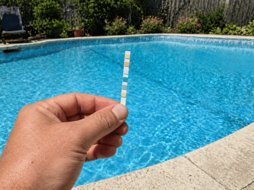 Hand holding a pool test strip in front of a clear swimming pool