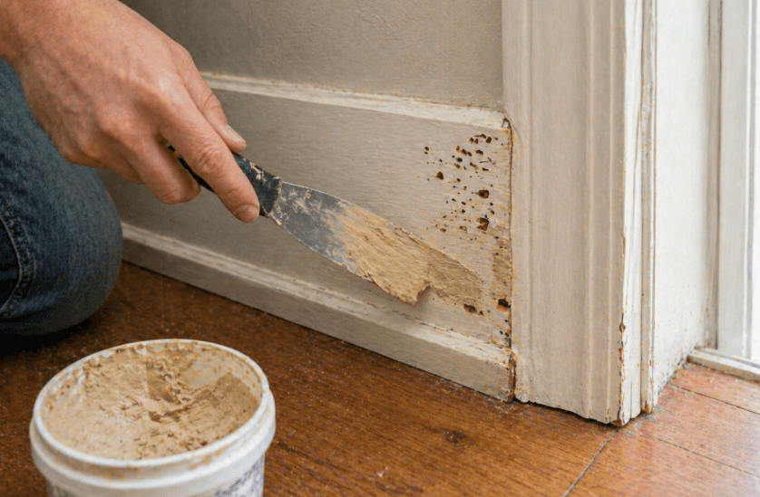 person applying wood filler to repair termite-damaged baseboard near door frame